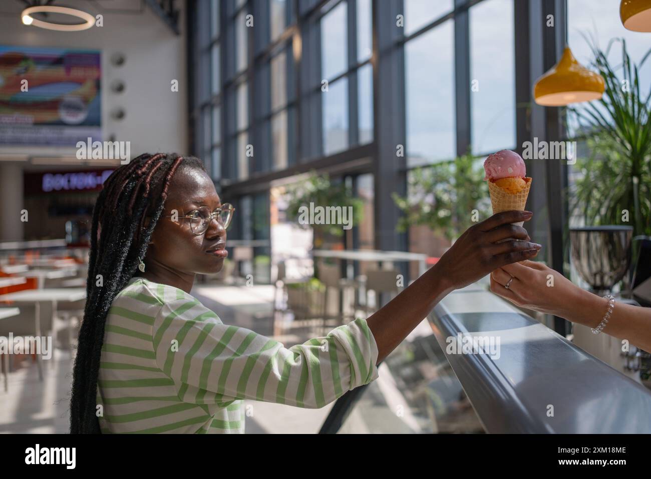 Junge afroamerikanische Frau mit Gläsern und Dreadlocks, die in einem hellen, modernen Café mit großen Fenstern eine Eiskegel erhält. Black Girl kauft kalte Desserts im Einkaufszentrum. Stockfoto