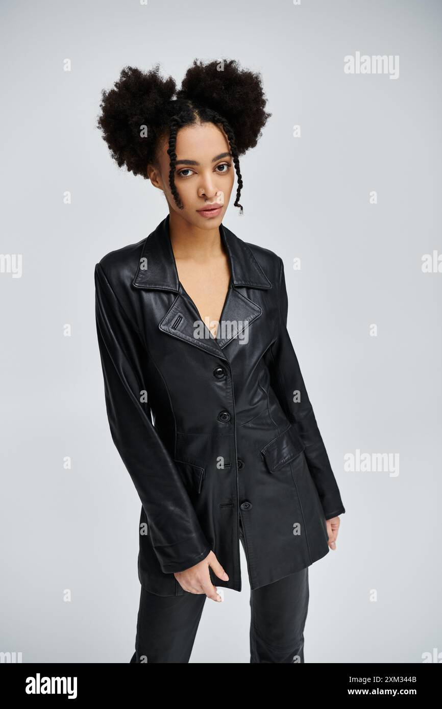 Eine junge afroamerikanische Frau in einem schwarzen Lederblazer posiert selbstbewusst vor einem grauen Hintergrund und zeigt einen stilvollen und modernen Look. Stockfoto
