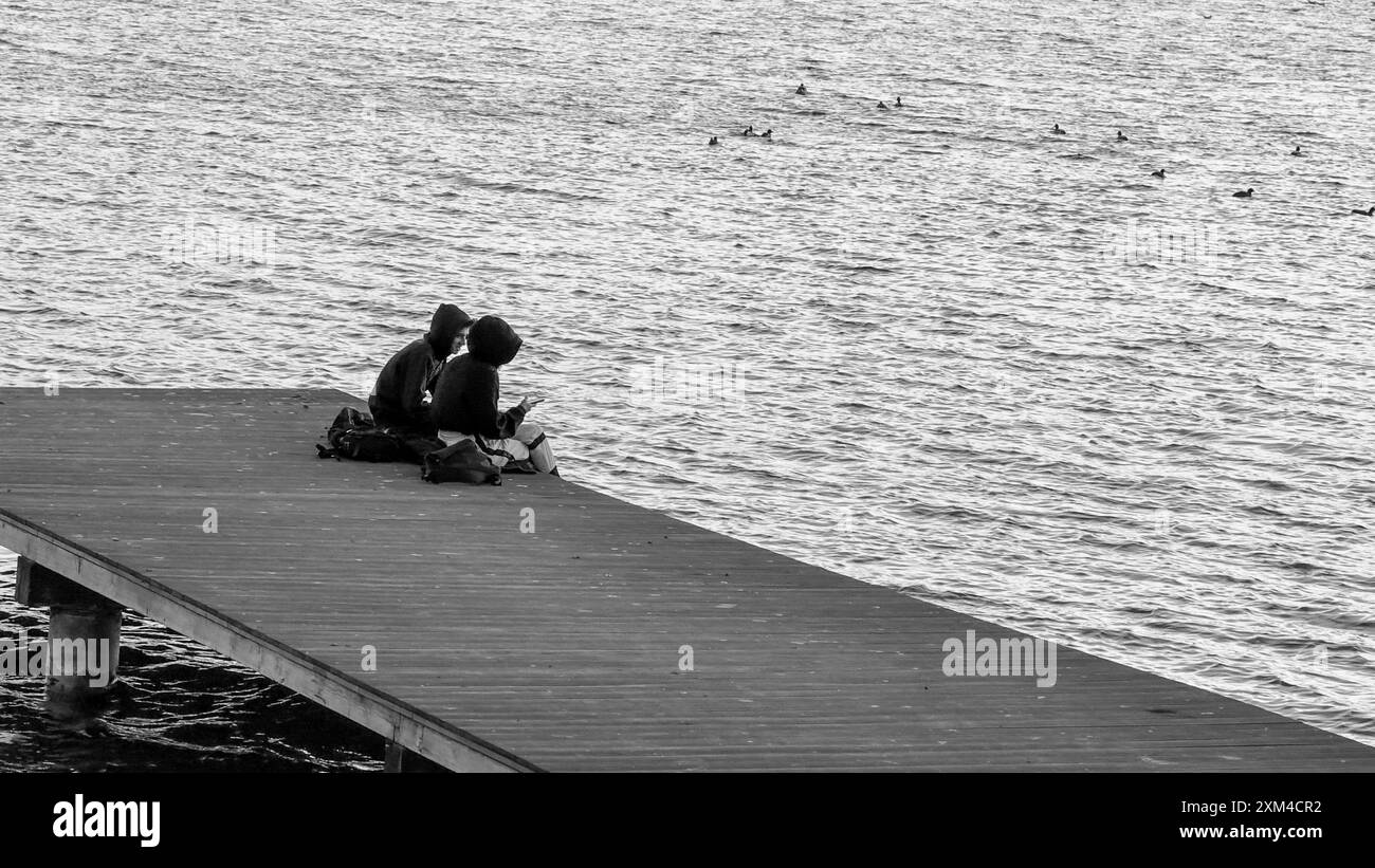 Zwei Personen sitzen auf einem Pier am Wasser, genießen einen ruhigen Moment mit Enten, die in der Nähe schwimmen, und fangen die Ruhe und Natur ein. Stockfoto