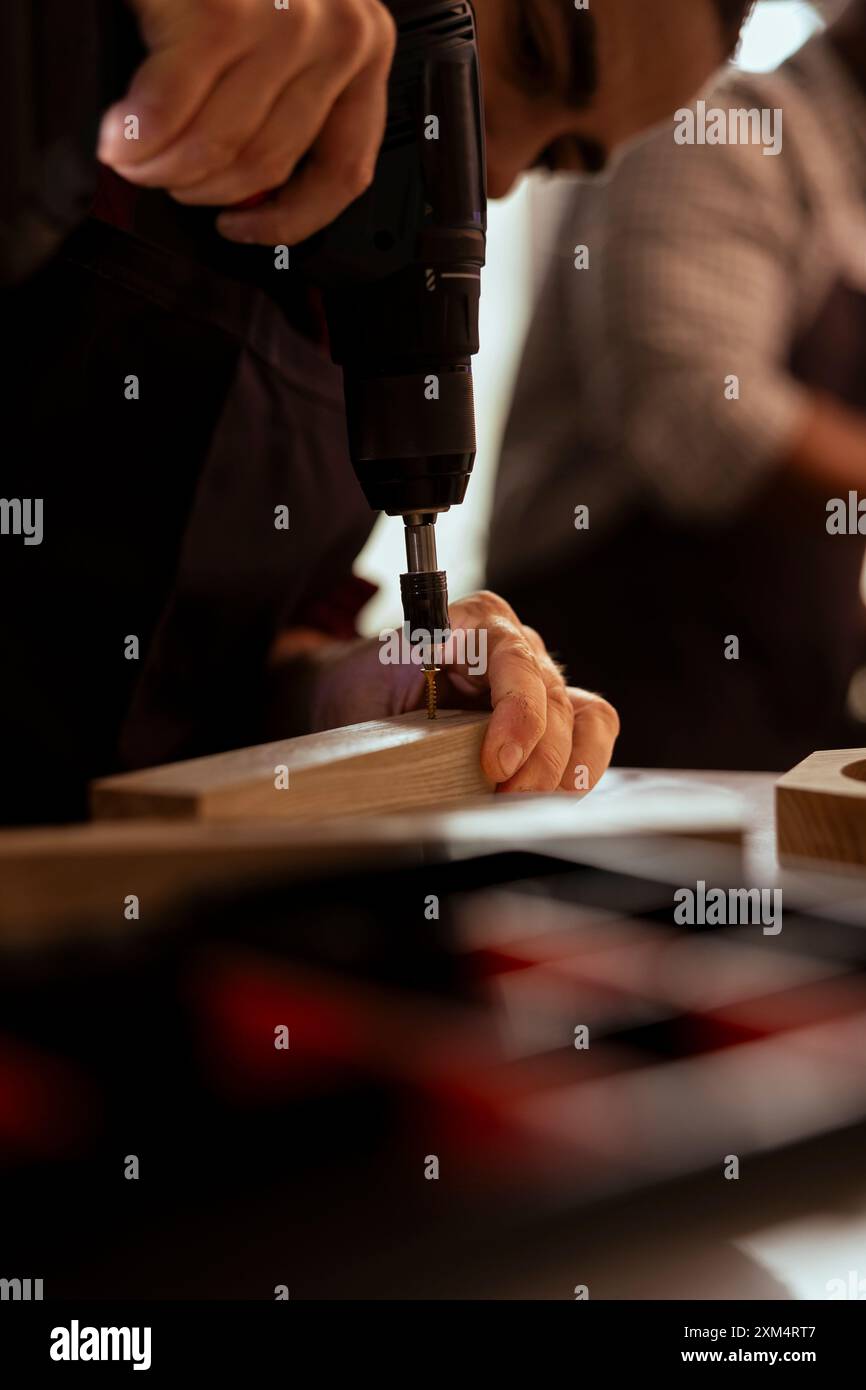 Zimmermann bohrt Loch in Holzblock im Studio, unterstützt von einem Kollegen, Nahaufnahme. Holzbearbeitungsexperte in der Tischlerei Nageleinbau in Holzstück mittels Bohrmaschine mit Lehrling im Hintergrund Stockfoto