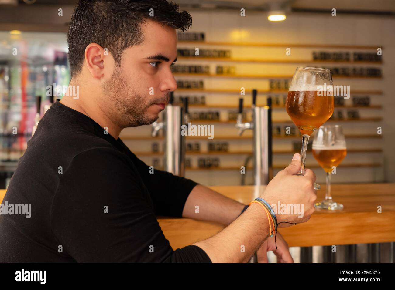 Ein junger Latino genießt ein Craft-Bier in einem Pub. Er beobachtet sorgfältig die Farbe und den Hopfen in dem Lager, das er verkostet. Stockfoto