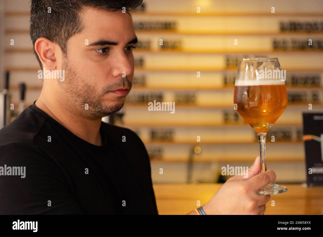 Ein junger Latino genießt ein Craft-Bier in einem Pub. Er beobachtet sorgfältig die Farbe und den Hopfen in dem Lager, das er verkostet. Stockfoto