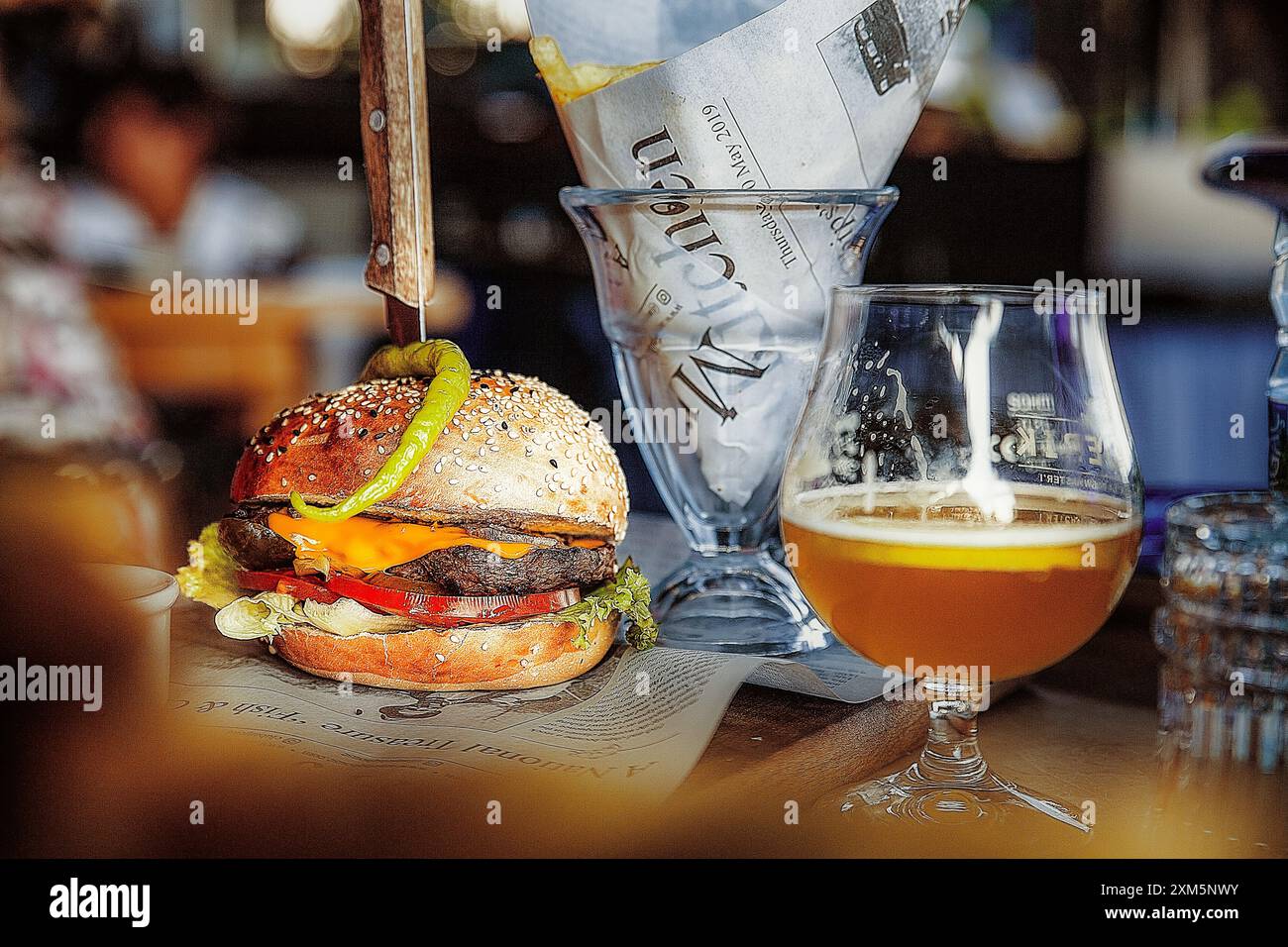 Ein saftiger Burger mit Patty, Käse und grüner Paprika mit einem Messer und einem Glas kaltes Bier mit Zeitungsdekorationen Stockfoto