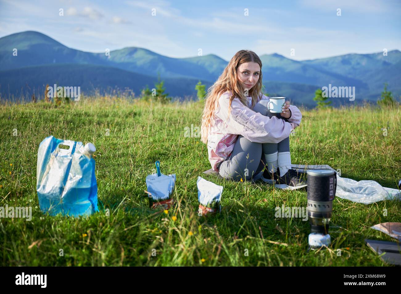 Die Touristin sitzt auf einem grasbewachsenen Hügel und hält den Becher. Reisender mit langen blonden Haaren. In der Nähe tragbarer Campingkocher, Essensbeutel, blaue Tasche. Die malerische Bergkulisse sorgt für eine ruhige Atmosphäre im Freien. Stockfoto