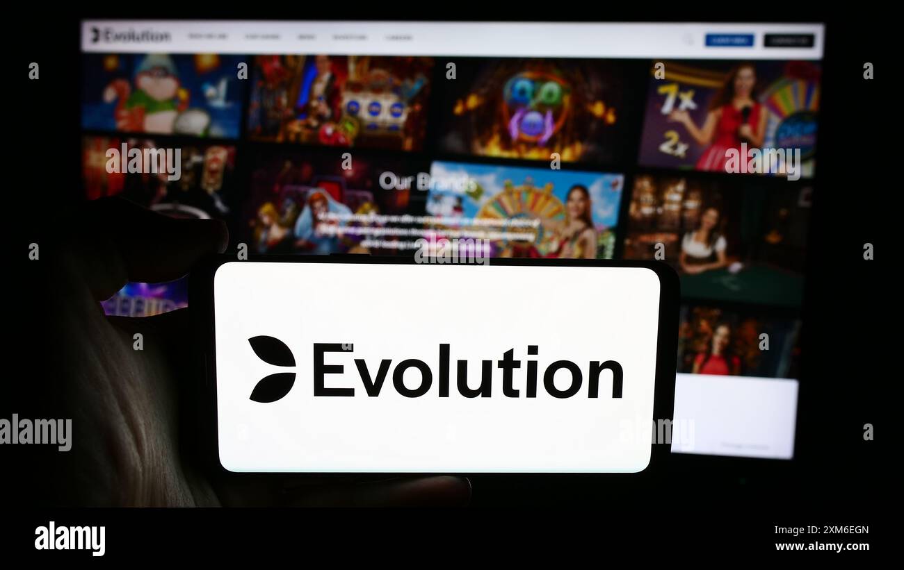 Person, die ein Mobiltelefon mit dem Logo des schwedischen Glücksspieltechnologieunternehmens Evolution ab vor der Webseite hält. Schwerpunkt auf dem Display des Telefons. Stockfoto