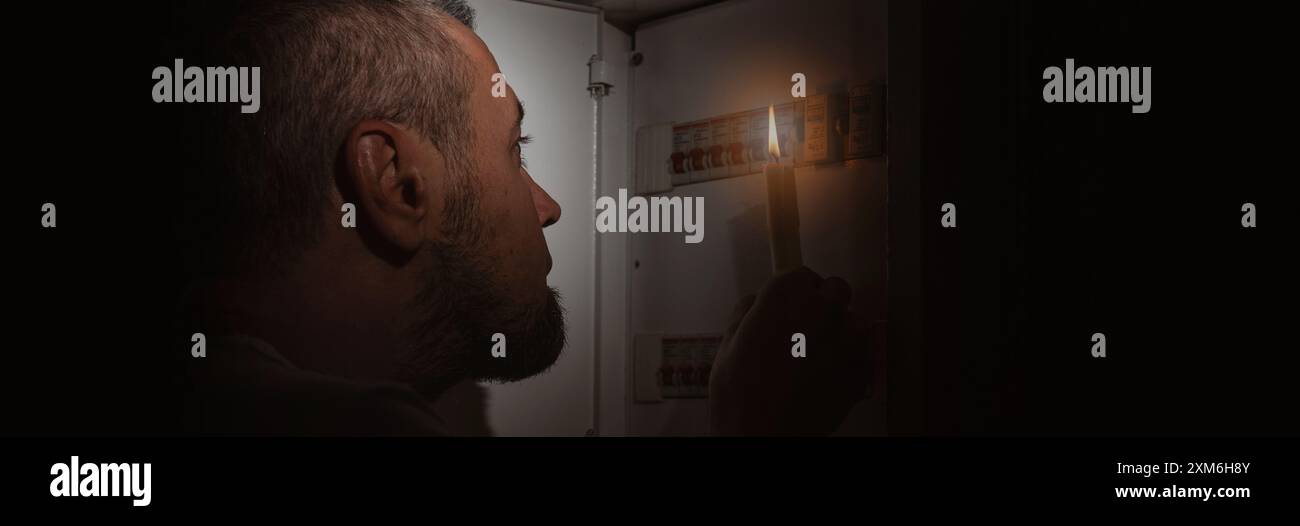 Blackout-Konzept. Ein verängstigter Mann mit einer Kerze während des Blackouts. Mann, der mit einer Kerze im Dunkeln in die Kamera schaut. Stockfoto