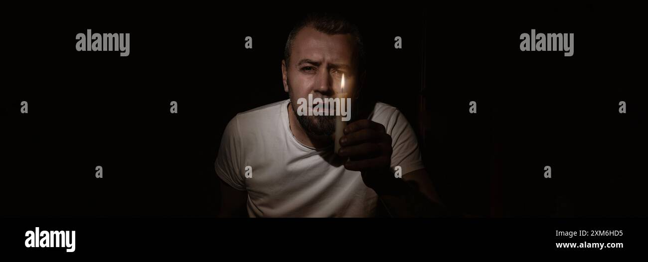 Blackout-Konzept. Ein verängstigter Mann mit einer Kerze während des Blackouts. Mann, der mit einer Kerze im Dunkeln in die Kamera schaut. Stockfoto