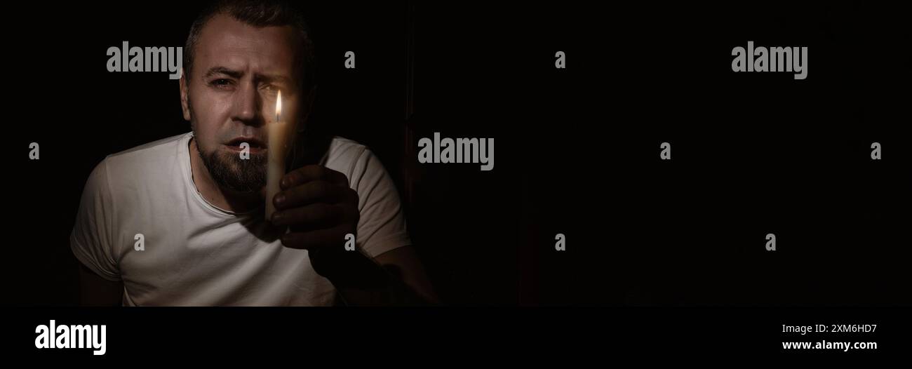 Blackout-Konzept. Ein verängstigter Mann mit einer Kerze während des Blackouts. Mann, der mit einer Kerze im Dunkeln in die Kamera schaut. Stockfoto
