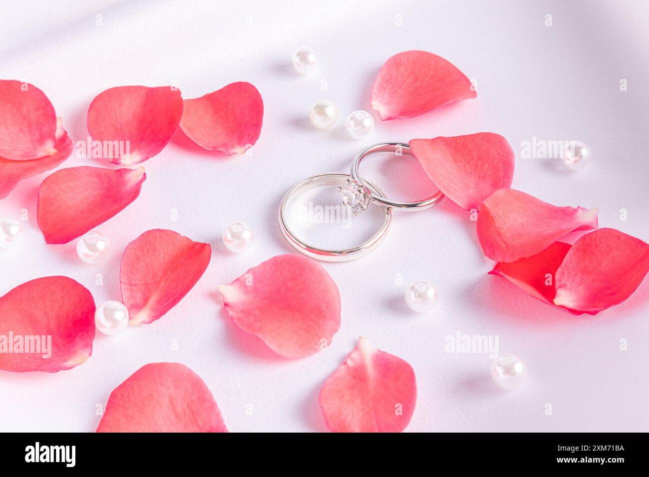 Zwei schicke Diamant-Eheringe aus Weißgold auf weißem Satin-Hintergrund mit Rosenblättern und Perlen. Draufsicht. Das Konzept der Hochzeit Stockfoto