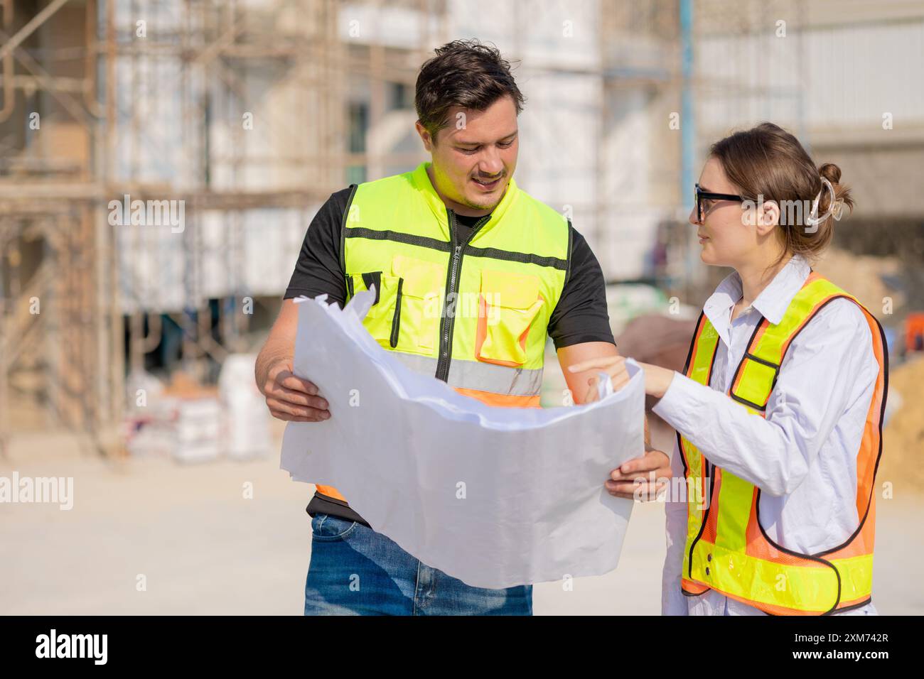 Zwei Bauarbeiter in Sicherheitswesten prüfen einen Plan auf einer Außenbaustelle, arbeiten zusammen und planen Arbeiten. Stockfoto