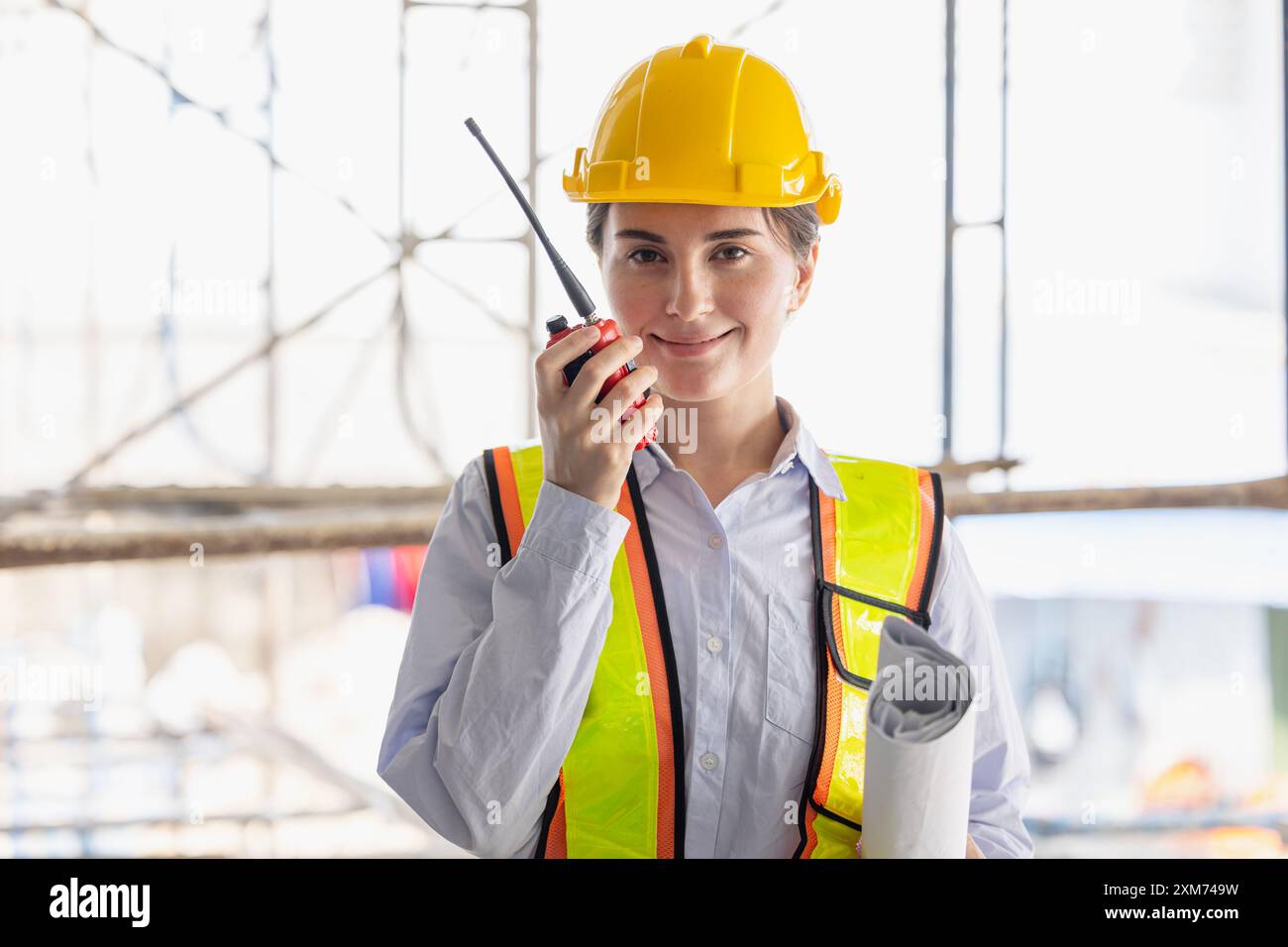 Weibliche Bauarbeiterin mit Schutzausrüstung und Schutzhelm, Kommunikation mit einem Funkgerät und Blaupausen auf einer Baustelle. Stockfoto