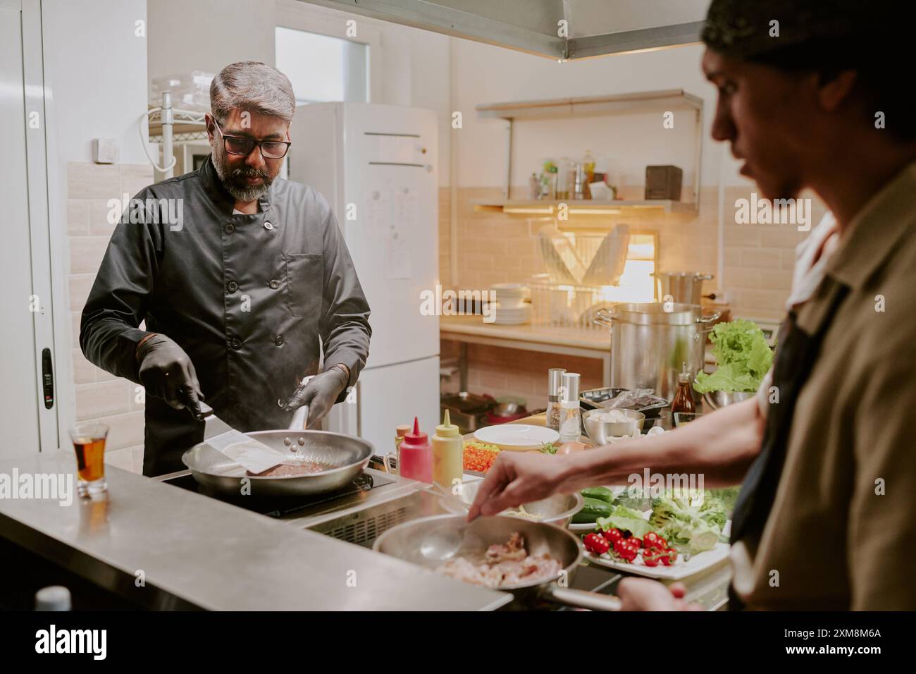 Professioneller indischer Koch in schwarzer Jacke und mit schwarzen Latexhandschuhen, der schwarzes angus-Steak anbrät, während sein Sous Chef Speck frittiert Stockfoto