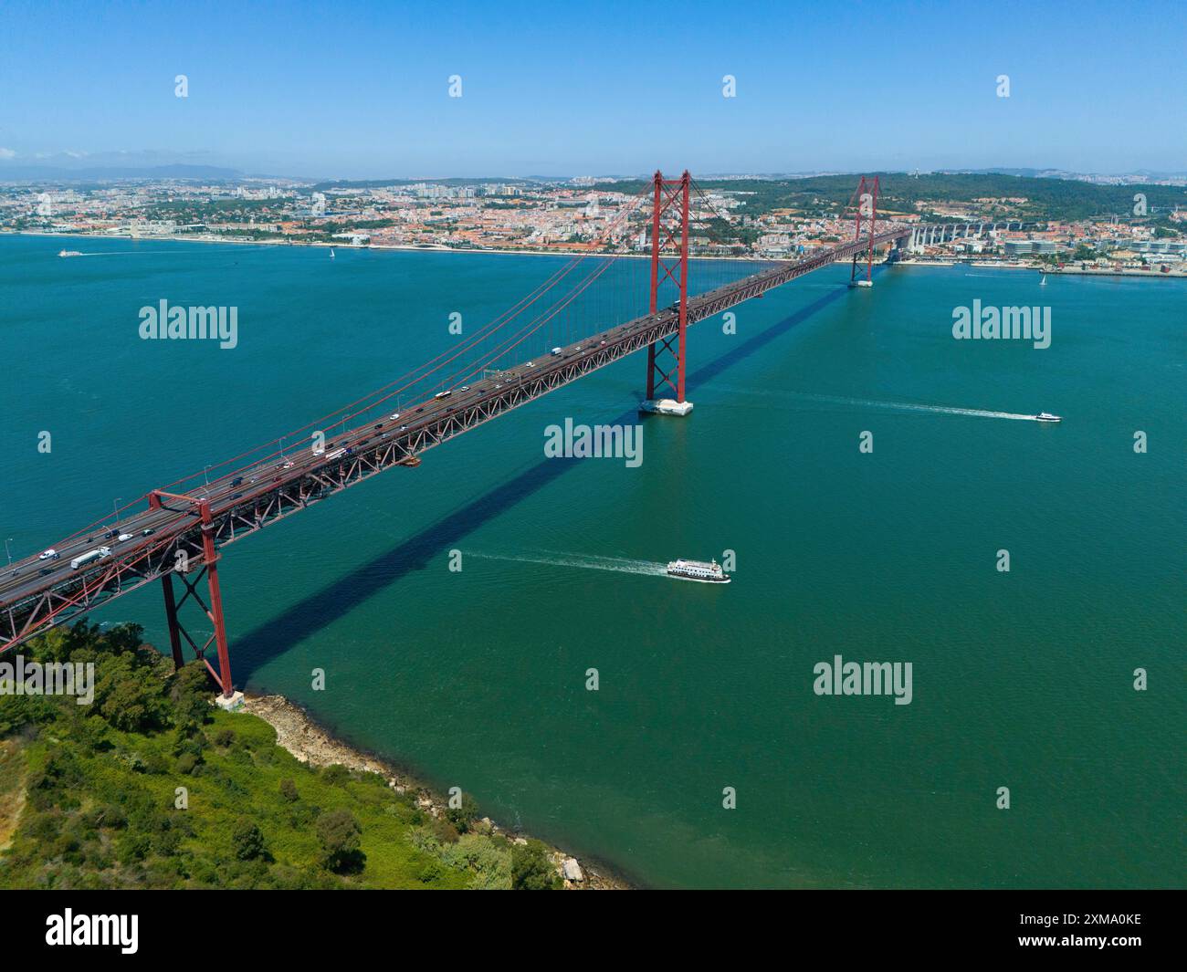 Rote Brücke überspannt das Wasser, gesäumt von Booten, mit einer Stadt und Natur im Hintergrund an einem sonnigen Tag, Blick aus der Luft, Ponte 25 de Abril, Brücke von Stockfoto