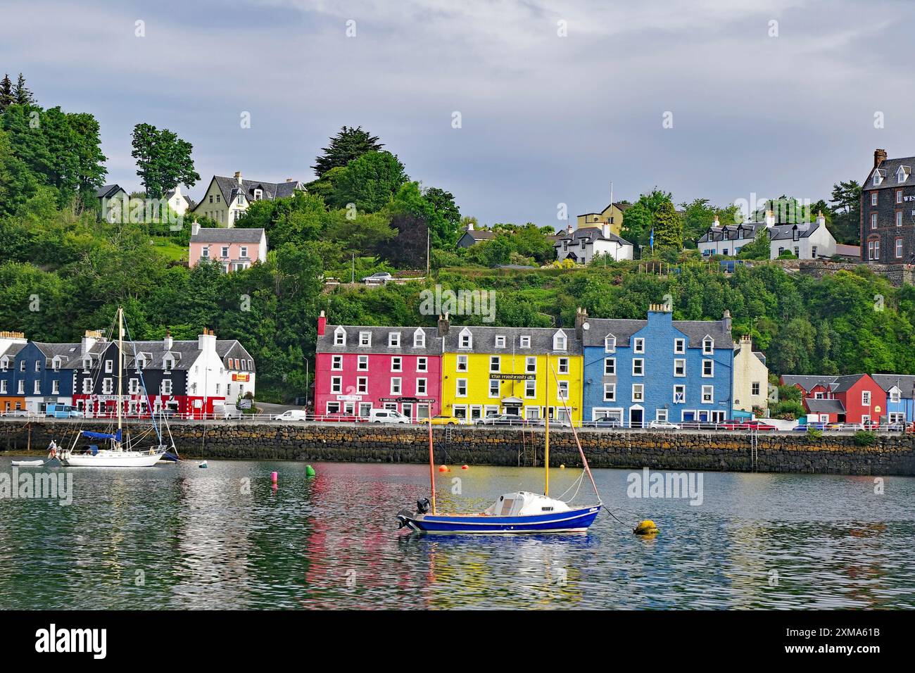 Häuser mit bunten Fassaden entlang eines Hafens, Boote im Vordergrund und ein grüner Hügel im Hintergrund unter einem blauen Himmel mit Wolken, Tobermory Stockfoto