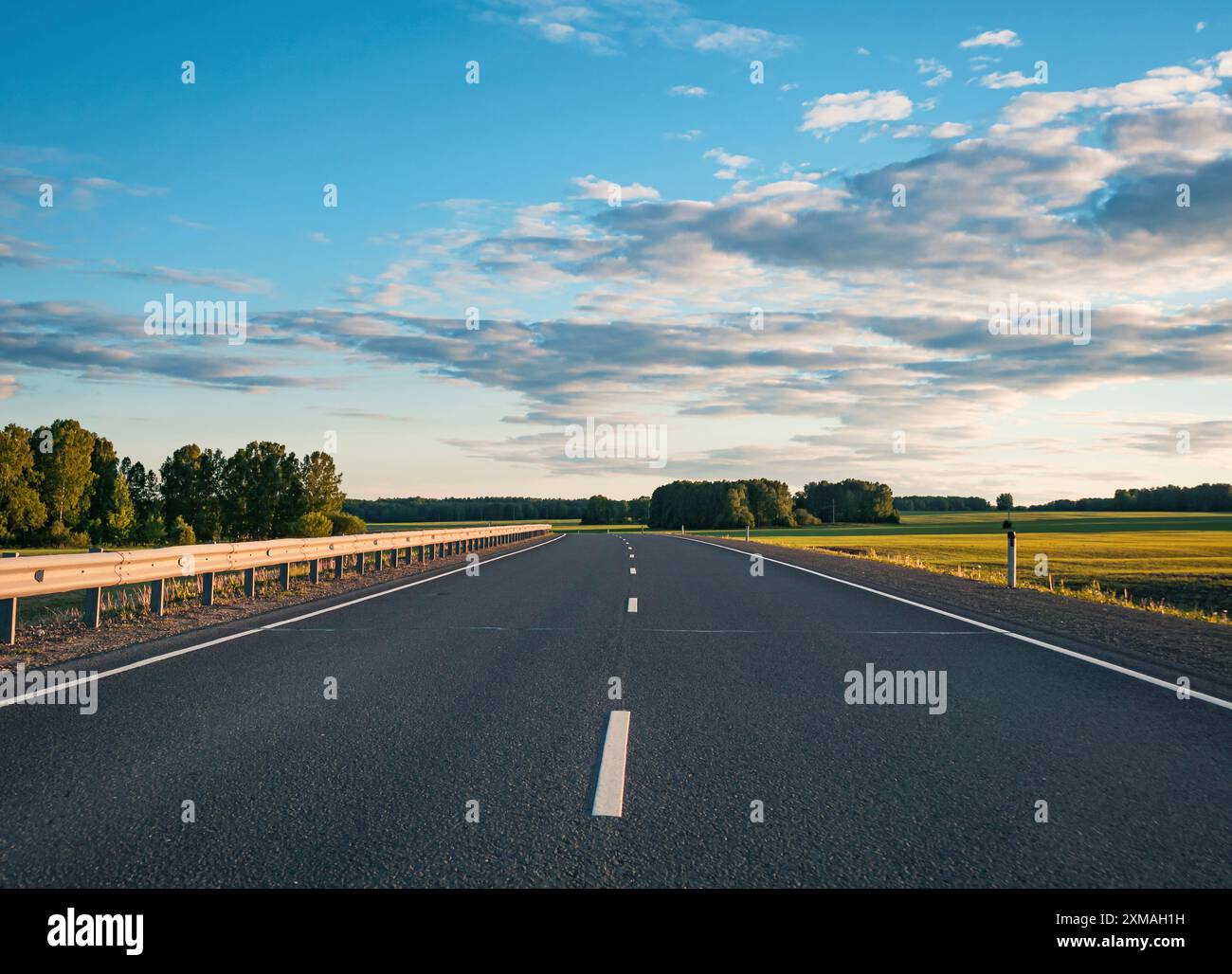 Endlose Asphaltstraße, die sich durch üppige grüne Felder unter offenem blauen Himmel erstreckt Stockfoto