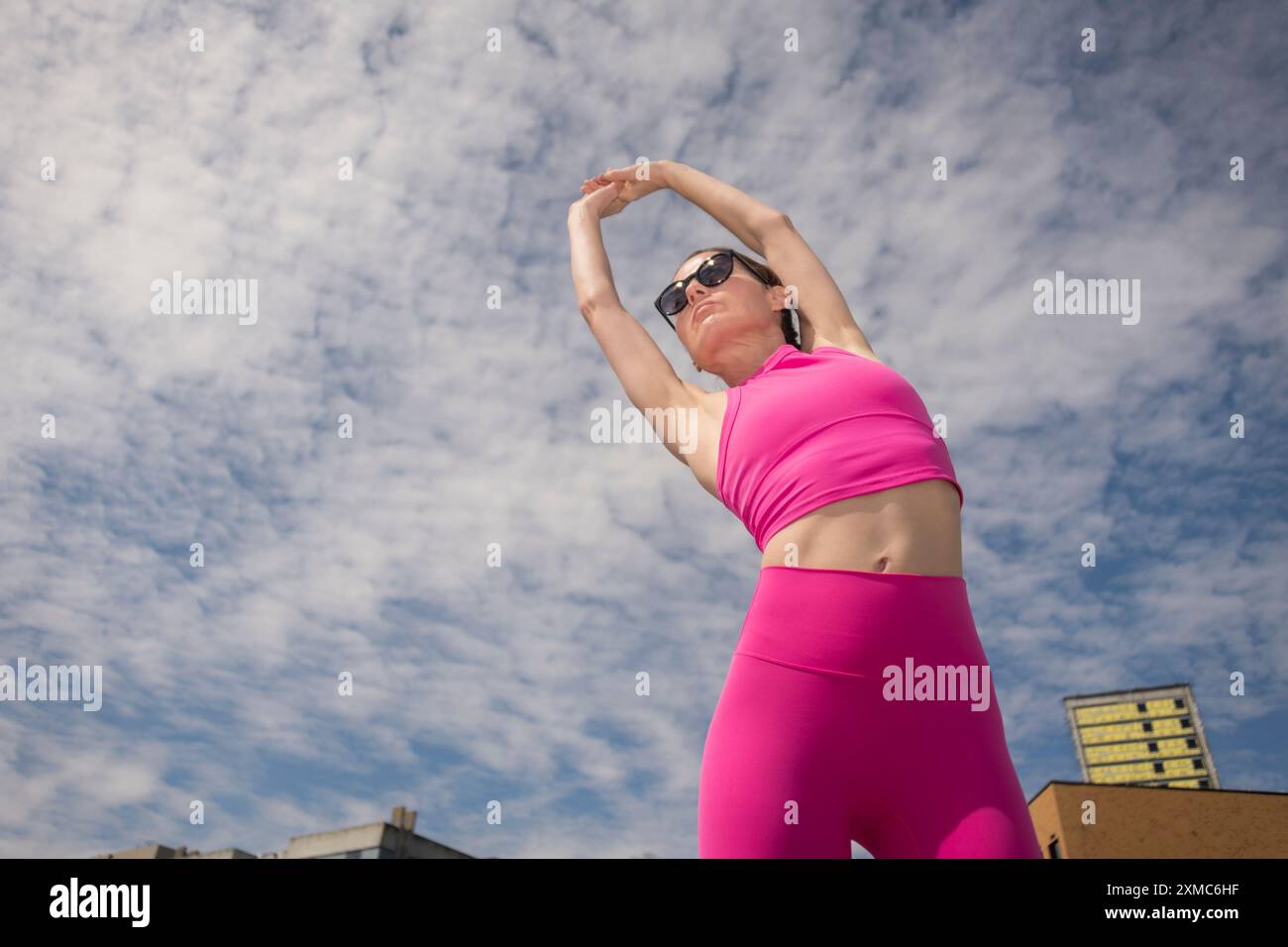 Sportliche Frau, die Arm-Dehnübungen in rosafarbener Sportkleidung macht, vor blauem Himmel Stockfoto