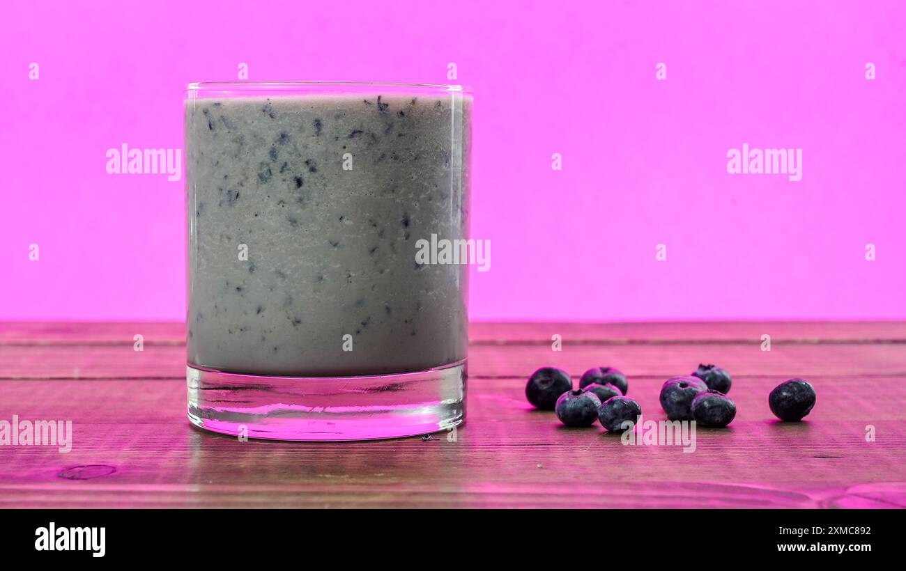 Neben frischen Blaubeeren wird ein leuchtender Heidelbeeren-Smoothie vor einem leuchtend rosa Hintergrund platziert Stockfoto