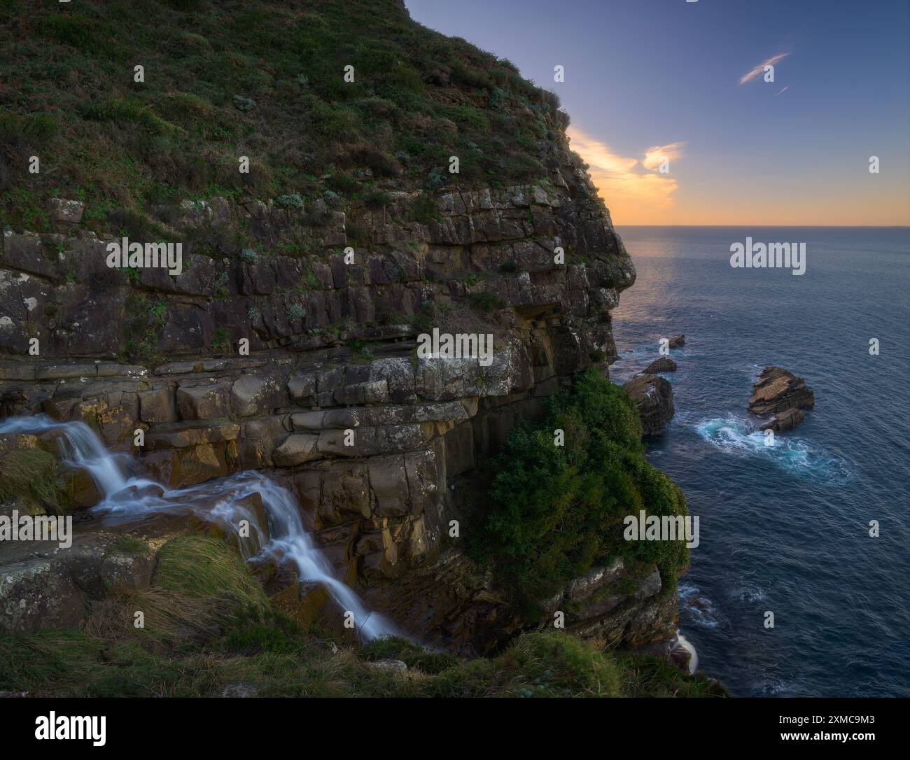 Der Bach stürzt ins Meer, eingerahmt von einer felsigen Küste. Im Schatten verborgener Vordergrund mit einem goldenen Sonnenuntergang, der den Hintergrund beleuchtet. Stockfoto