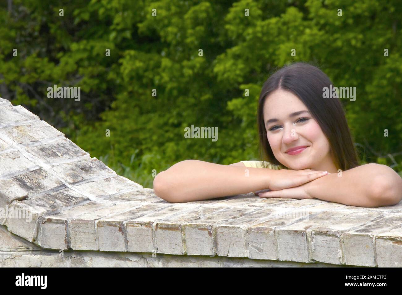 Weibliche Teenager legt ihre Ellbogen auf eine Ziegelwand, während sie hinüberblickt. Sie hat lange Haare und lächelt. Stockfoto