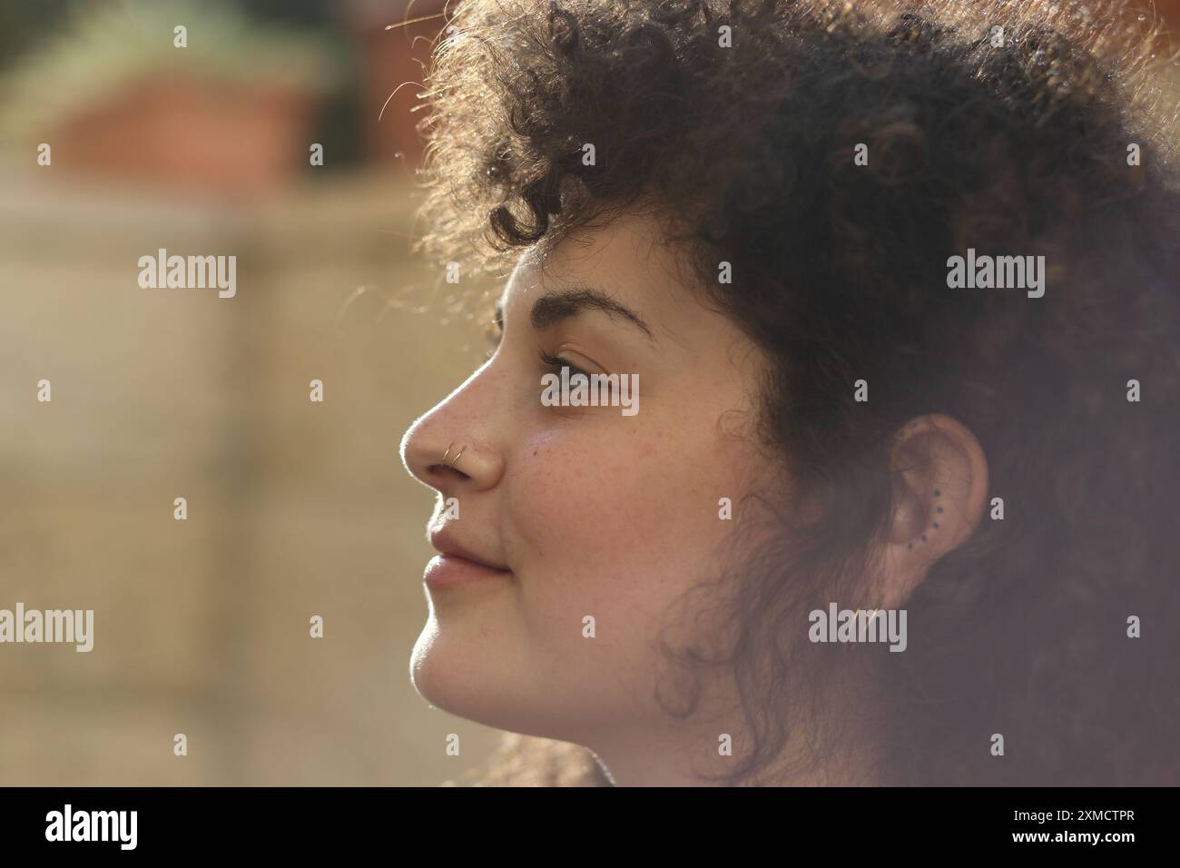 Profil einer schönen 24-jährigen Frau mit lockigem Haar, die Ohrringe und Nasenring trägt. Sie lächelt leicht, während die Sonne von hinten kommt und ein Aufleuchten erzeugt Stockfoto