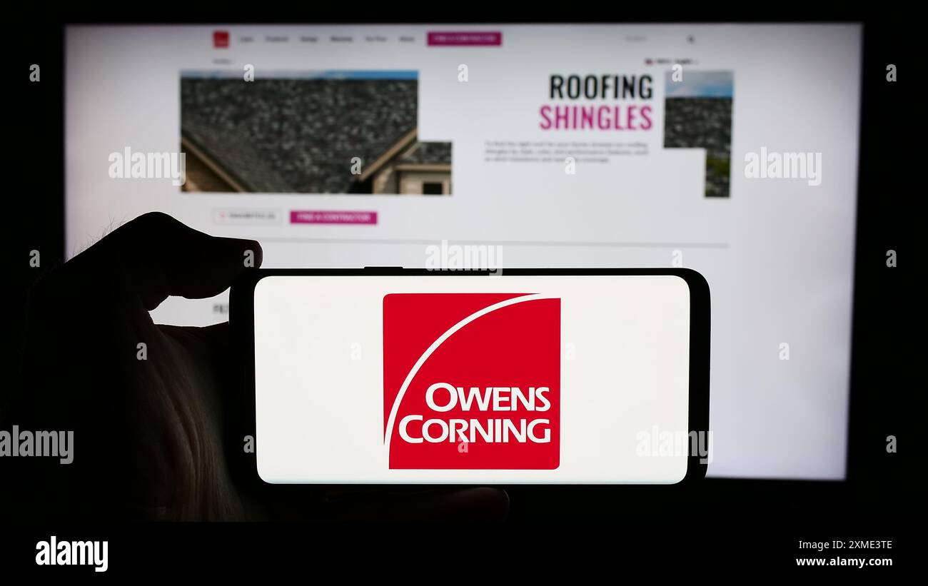 Person, die ein Mobiltelefon mit dem Logo des US-amerikanischen Baustoffunternehmens Owens Corning vor der Business-Webseite hält. Schwerpunkt auf dem Display des Telefons. Stockfoto