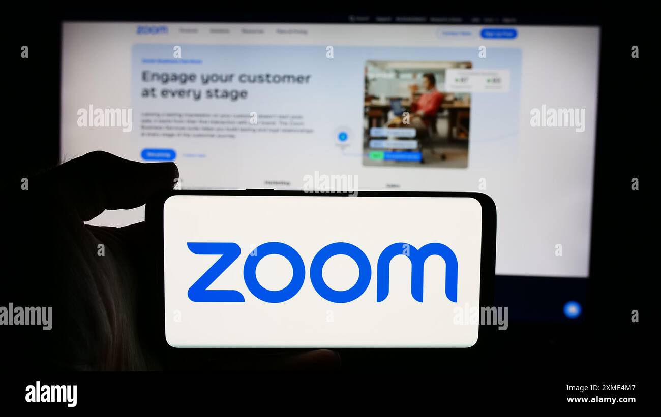 Person, die ein Mobiltelefon mit dem Logo des US-amerikanischen Kommunikationsunternehmens Zoom Video Communications Inc. Vor der Webseite hält. Schwerpunkt auf dem Display des Telefons. Stockfoto
