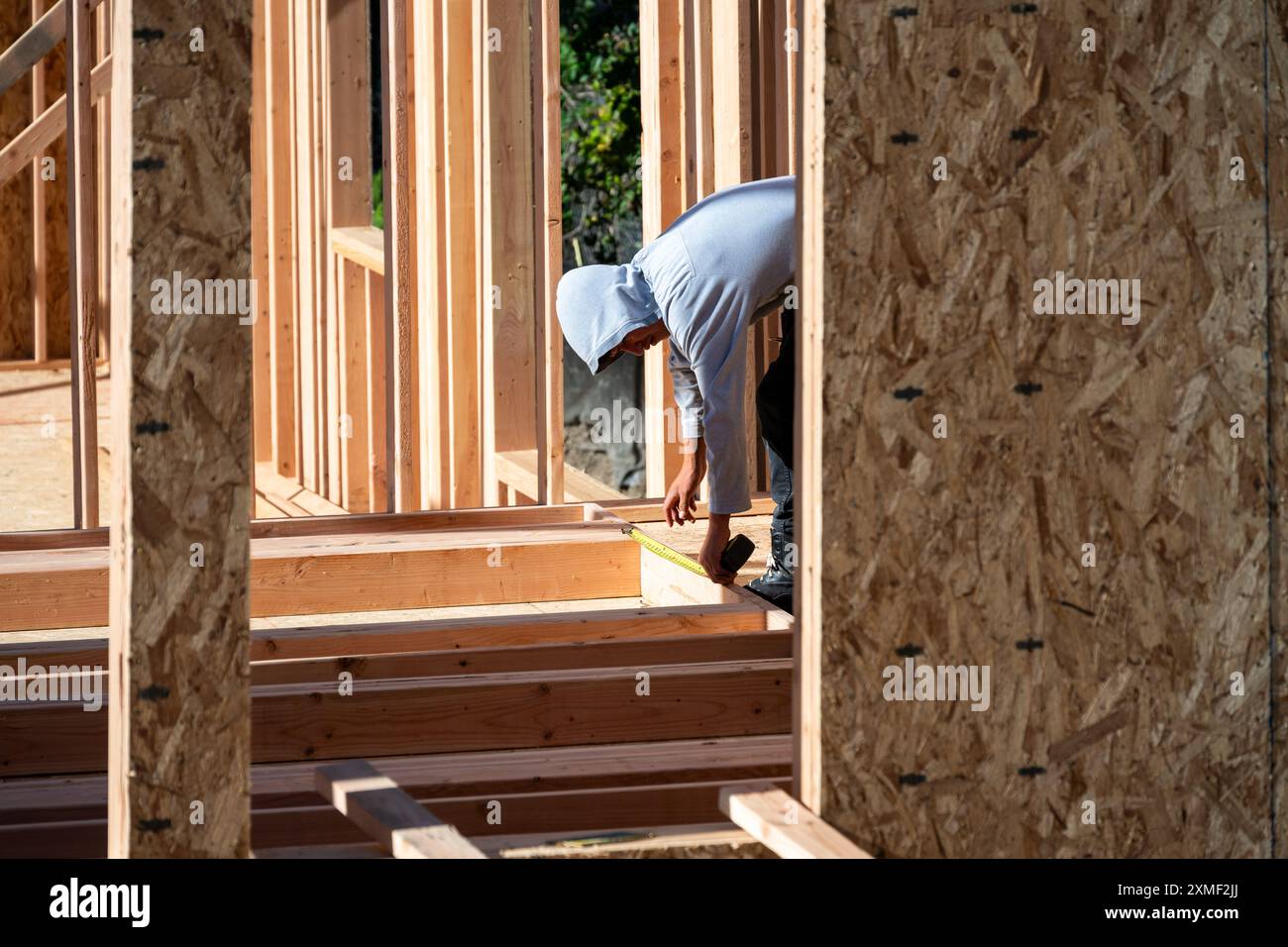 Bauarbeiter im Werkzeuggürtel, der mit einem Maßband an neuen Hausholzrahmen arbeitet, Sommerbausaison Stockfoto