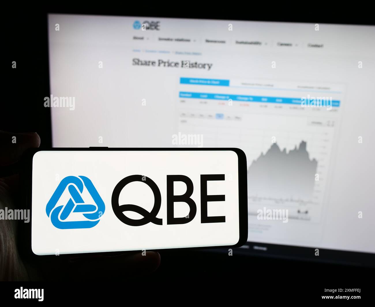 Person, die ein Mobiltelefon mit dem Logo der australischen QBE Insurance Group Limited vor der Webseite des Unternehmens hält. Schwerpunkt auf dem Display des Telefons. Stockfoto