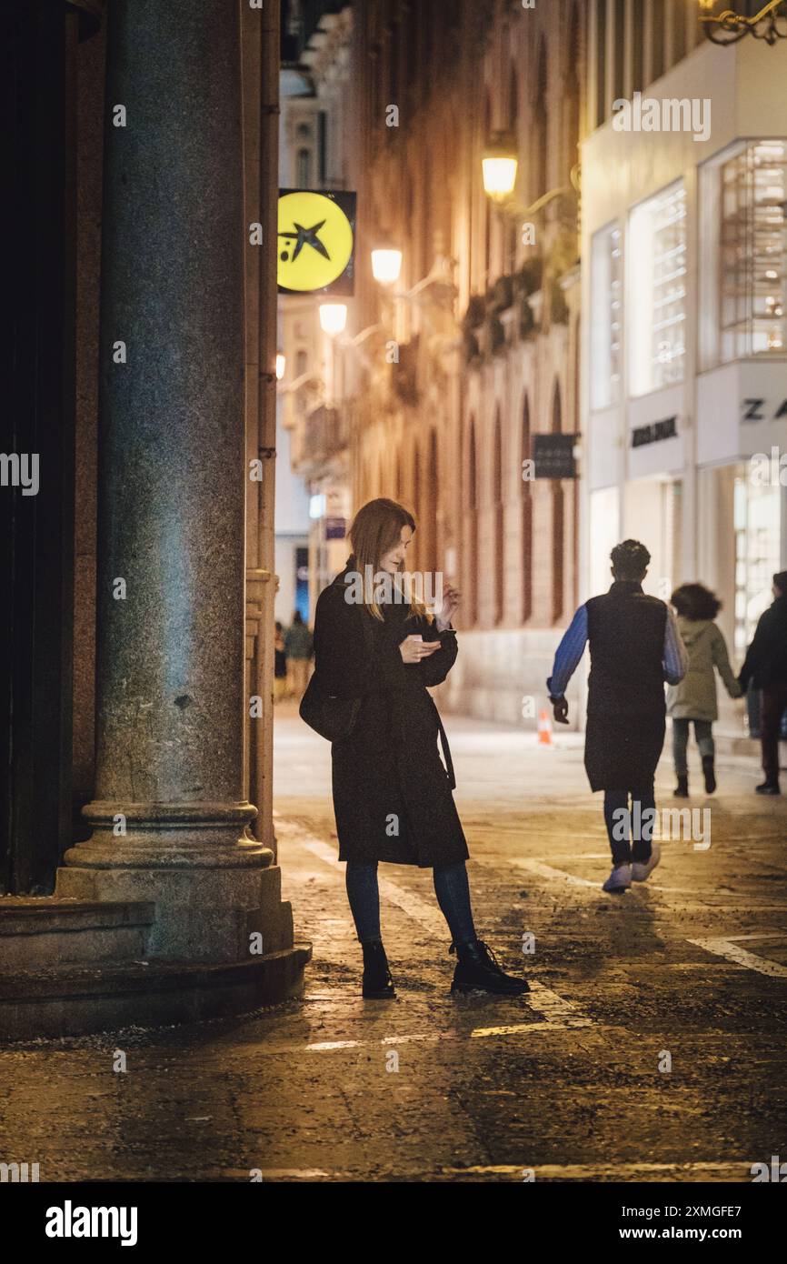 Städtische Nachtszene mit einer Frau, die Telefon benutzt, Fußgänger, Ladenfronten und beleuchtetem Kreuzungsschild. Stadtstraße mit Ambientebeleuchtung. Stockfoto