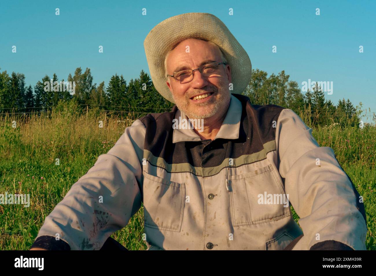 Ein Mann mit Imkerei-Anzug und Strohhut sitzt auf dem Gras auf einem sonnendurchfluteten Feld und genießt eine Pause. Die ruhige Landschaft und das helle Sonnenlicht sind in den Vordergrund gerückt Stockfoto
