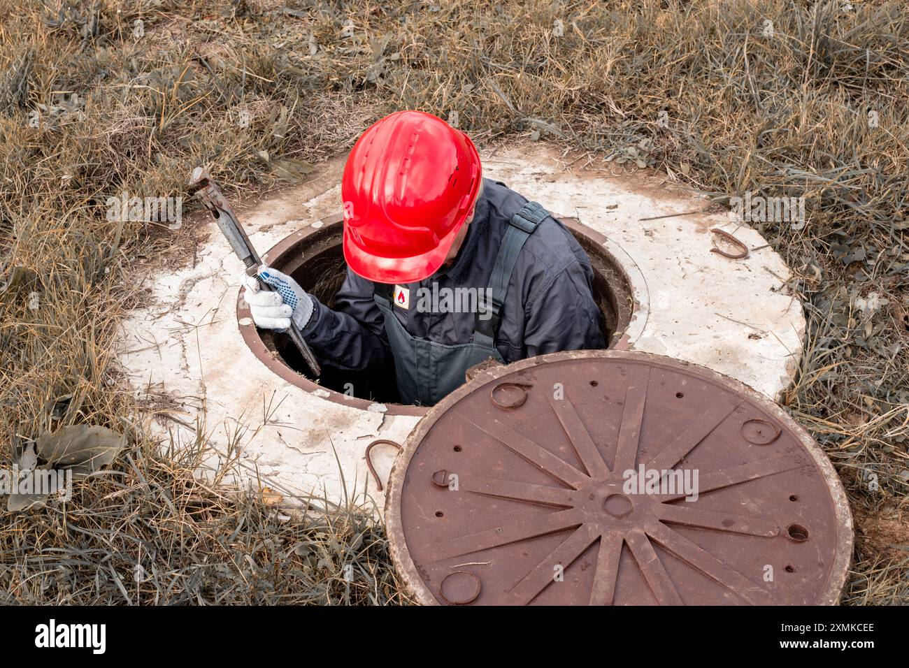 Ein Arbeiter in einem Helm sitzt mit einem verstellbaren Schraubenschlüssel in einem Wasserbrunnen. Wartung und Reparatur der Wasserversorgungseinheit. Stockfoto