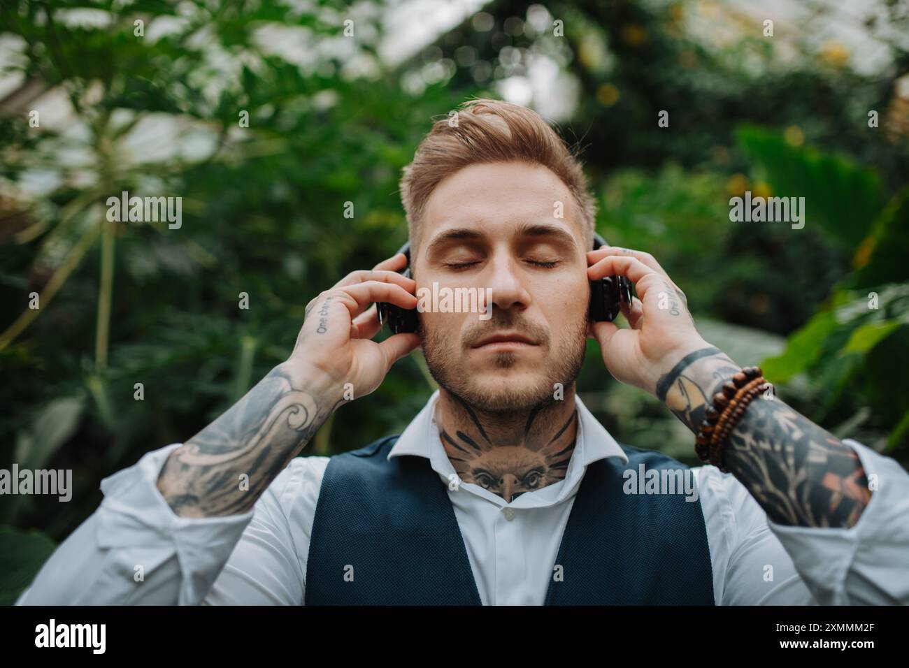 Gutaussehender Mann mit Tätowierung, der Meditationsmusik im botanischen Garten zuhört, inmitten von üppig grünem Laub. Er kümmert sich um seine psychische Gesundheit Stockfoto