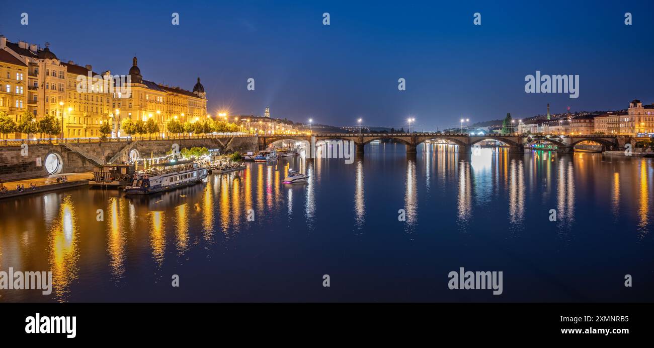 Blick auf den Rasin-Embankment und die Palacky-Brücke in Prag, Tschechien in der Abenddämmerung. Die Brücke ist beleuchtet und die Lichter der Stadt spiegeln sich im Wasser. Es gibt Boote auf dem Fluss, und der Himmel ist tief blau. Stockfoto