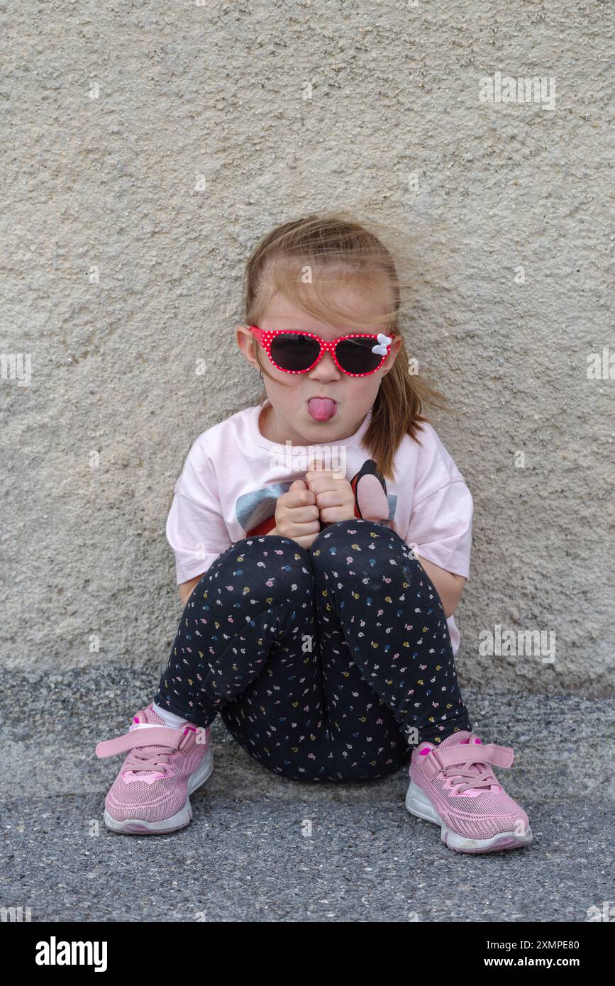 Das kleine kaukasische Mädchen sitzt an einer Wand, die die Zunge ausstreckt Stockfoto