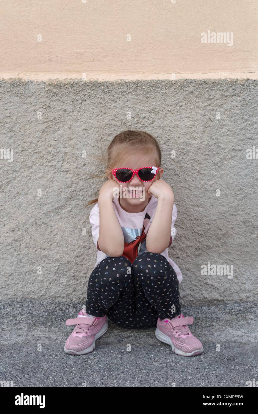 Das kleine kaukasische Mädchen sitzt mit Sonnenbrille an der Wand Stockfoto