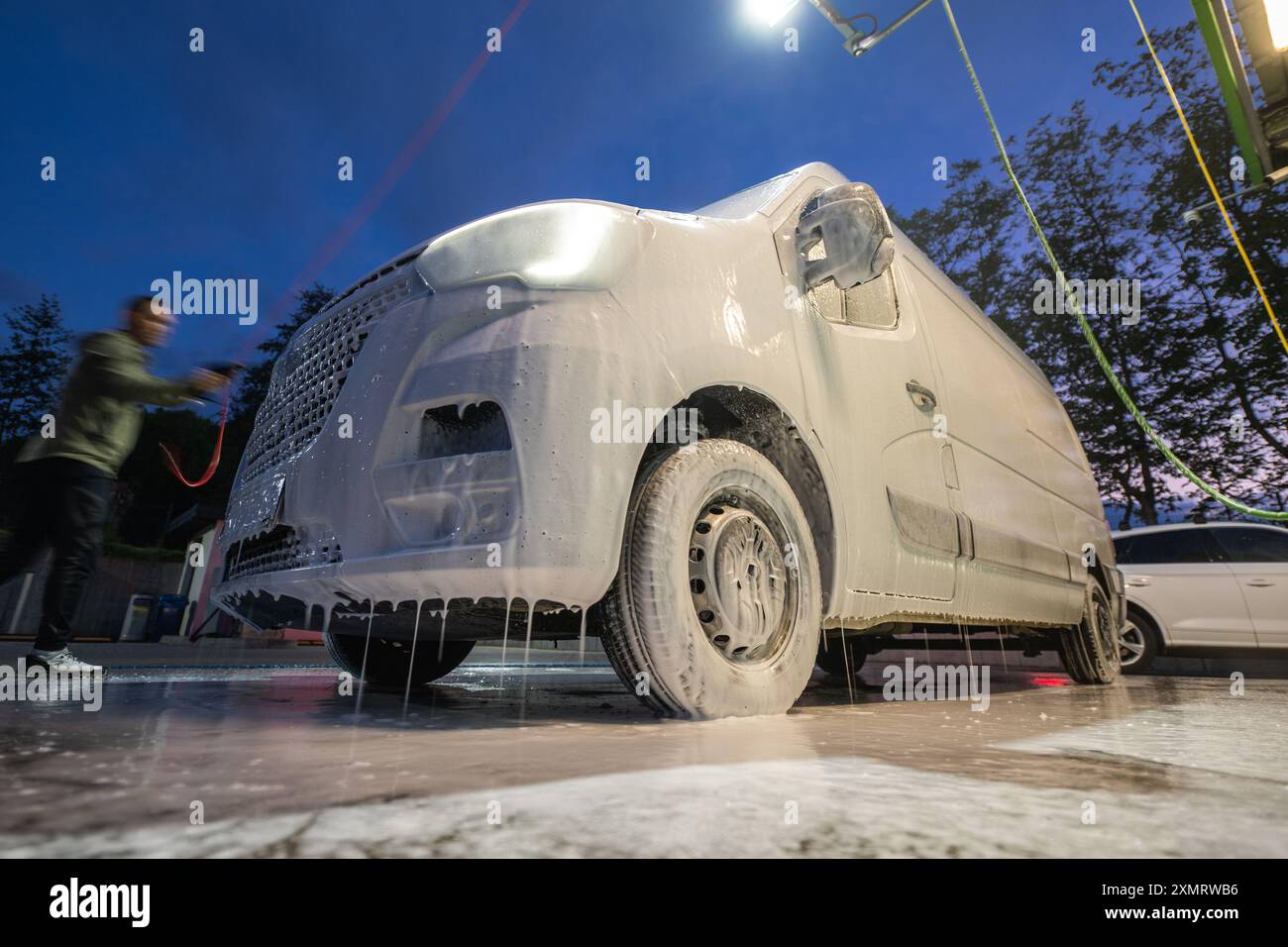 Ein Arbeiter trägt abends dicken Schaum auf einen Van an einer Autowaschanlage auf. Das Fahrzeug, teilweise mit Schaumstoff bedeckt, wird durch Deckenleuchten beleuchtet, wodurch entsteht Stockfoto