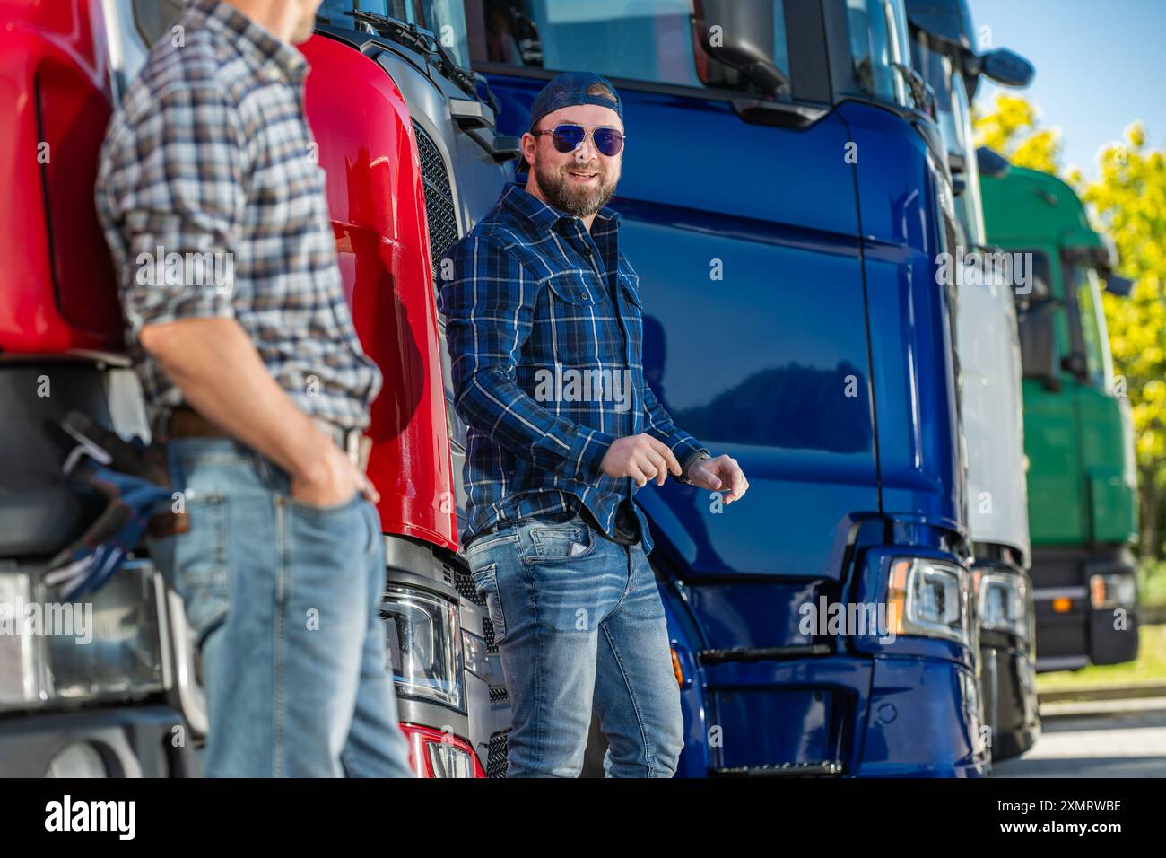 Zwei Männer in lässiger Kleidung unterhalten sich bei einer Reihe bunter Lastwagen auf einem Transporthof. Die helle Sonne wirft ein warmes Licht, während sie sich in Conversati einmischen Stockfoto
