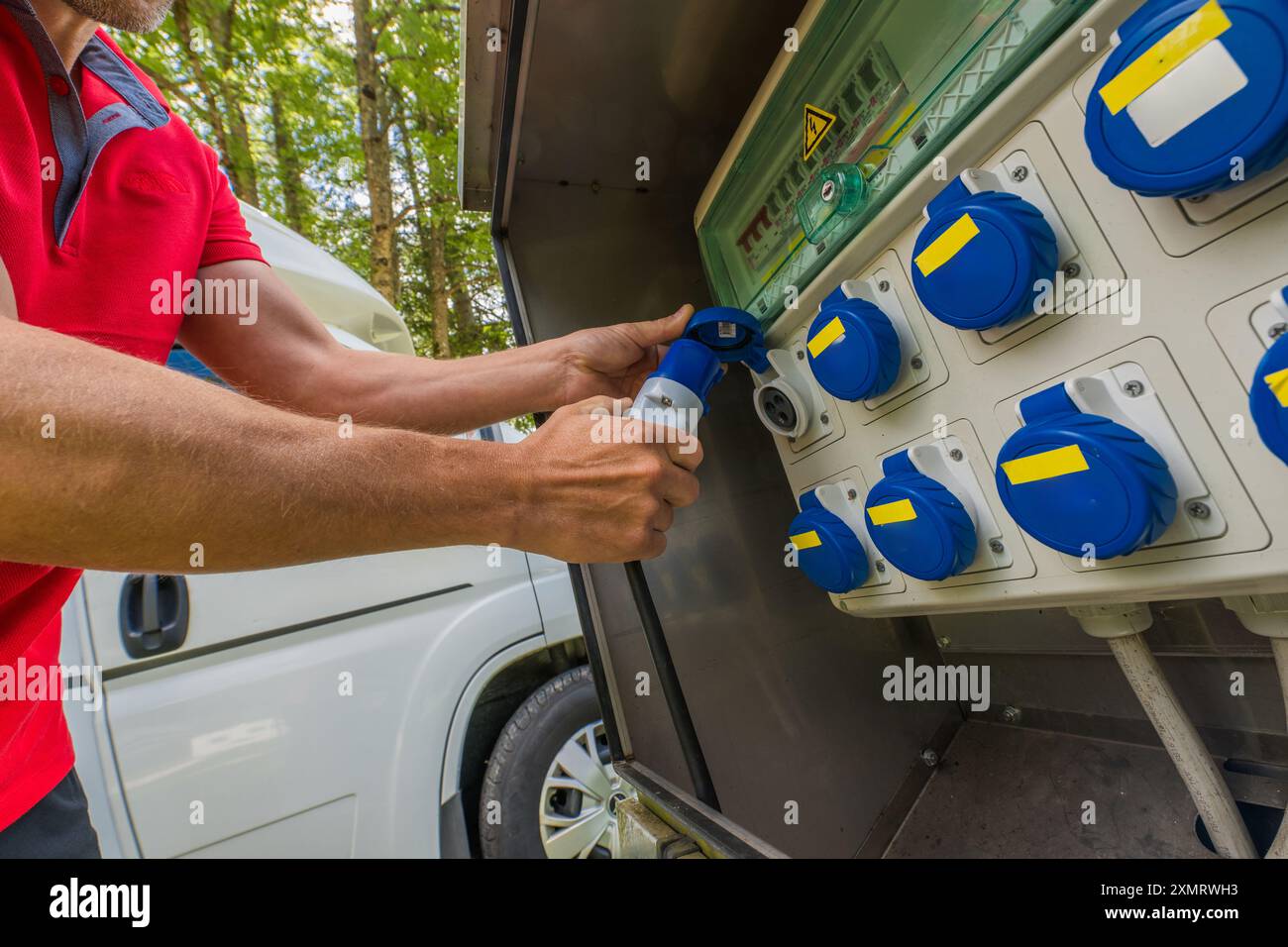 Eine Person schließt ein Netzkabel an eine Versorgungsbox auf einem von Bäumen umgebenen Campingplatz an. Die Szene erfasst die Vorbereitung auf den Stromeingang Stockfoto