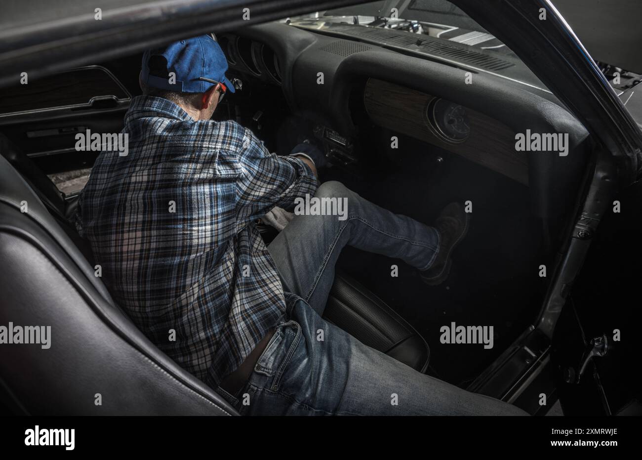 Ein junger Mechaniker kniet in einem Oldtimer nieder und arbeitet in einer gut beleuchteten Werkstatt intensiv am Armaturenbrett. Die Werkzeuge sind verstreut und spiegeln das Leben wider Stockfoto