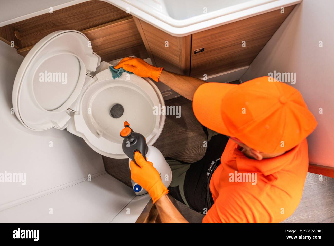 Ein professioneller Reiniger mit orangefarbener Kappe und Handschuhen reinigt aktiv eine weiße Toilette in einem modernen Badezimmer. Der Reiniger wendet eine Desinf an Stockfoto