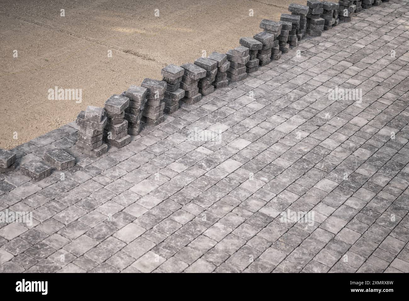 Eine ordentliche Anordnung frisch gelegter Kopfsteinpflaster erstreckt sich über den Vordergrund, während ordentlich gestapelte Pflastersteine neben einem Sandbereich sitzen. Diese Messung Stockfoto