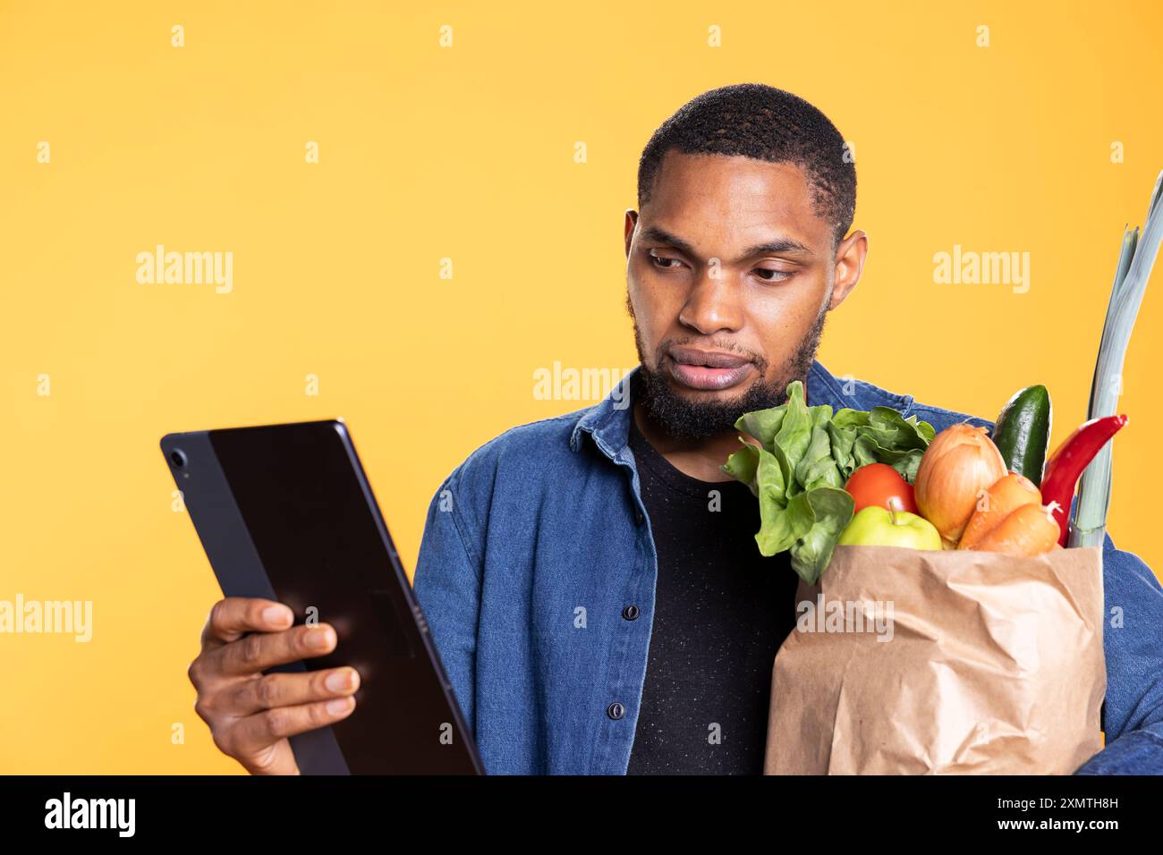 Ein afroamerikaner schaut seine Social-Media-Apps im Studio und trägt eine Papiertüte voller Lebensmittel aus dem Supermarkt. Glücklicher, entspannter Mann, der Spaß beim Surfen im Internet hatte, umweltfreundlich. Stockfoto