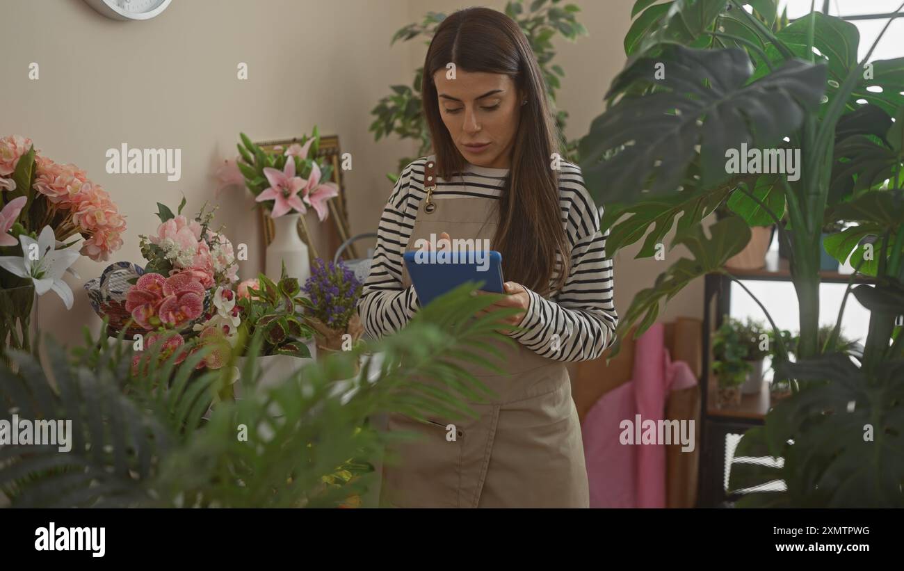Eine junge hispanische Frau steht drinnen zwischen Pflanzen, konzentriert auf ihr Tablet, während sie von einem gemütlichen Raum umgeben ist. Stockfoto