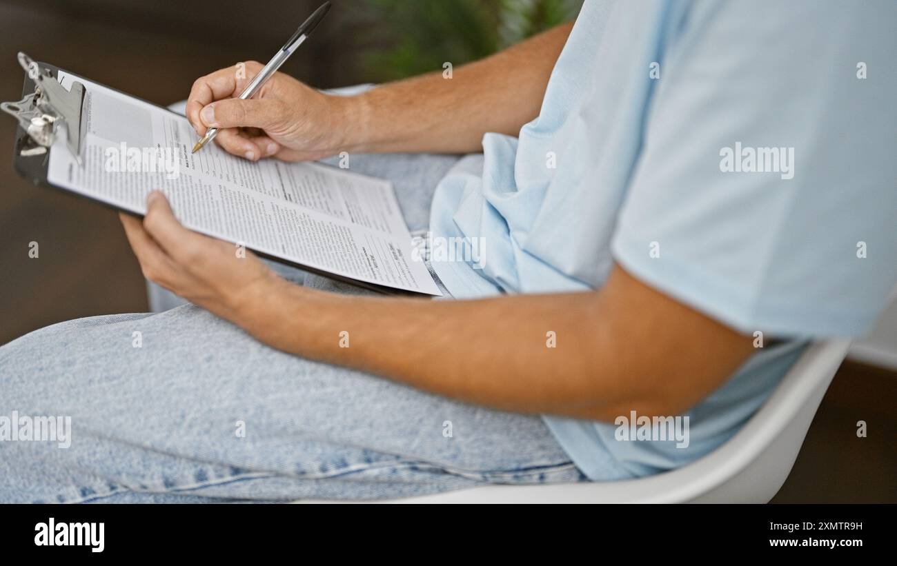 Hispanischer Mann in lässiger Kleidung sitzt drinnen und füllt Papiere auf einem Klemmbrett aus. Stockfoto