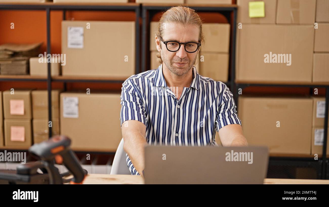 Ein fokussierter Mann mit Brille und gestreiftem Hemd, der an einem Laptop in einem Lagerhaus arbeitet. Stockfoto