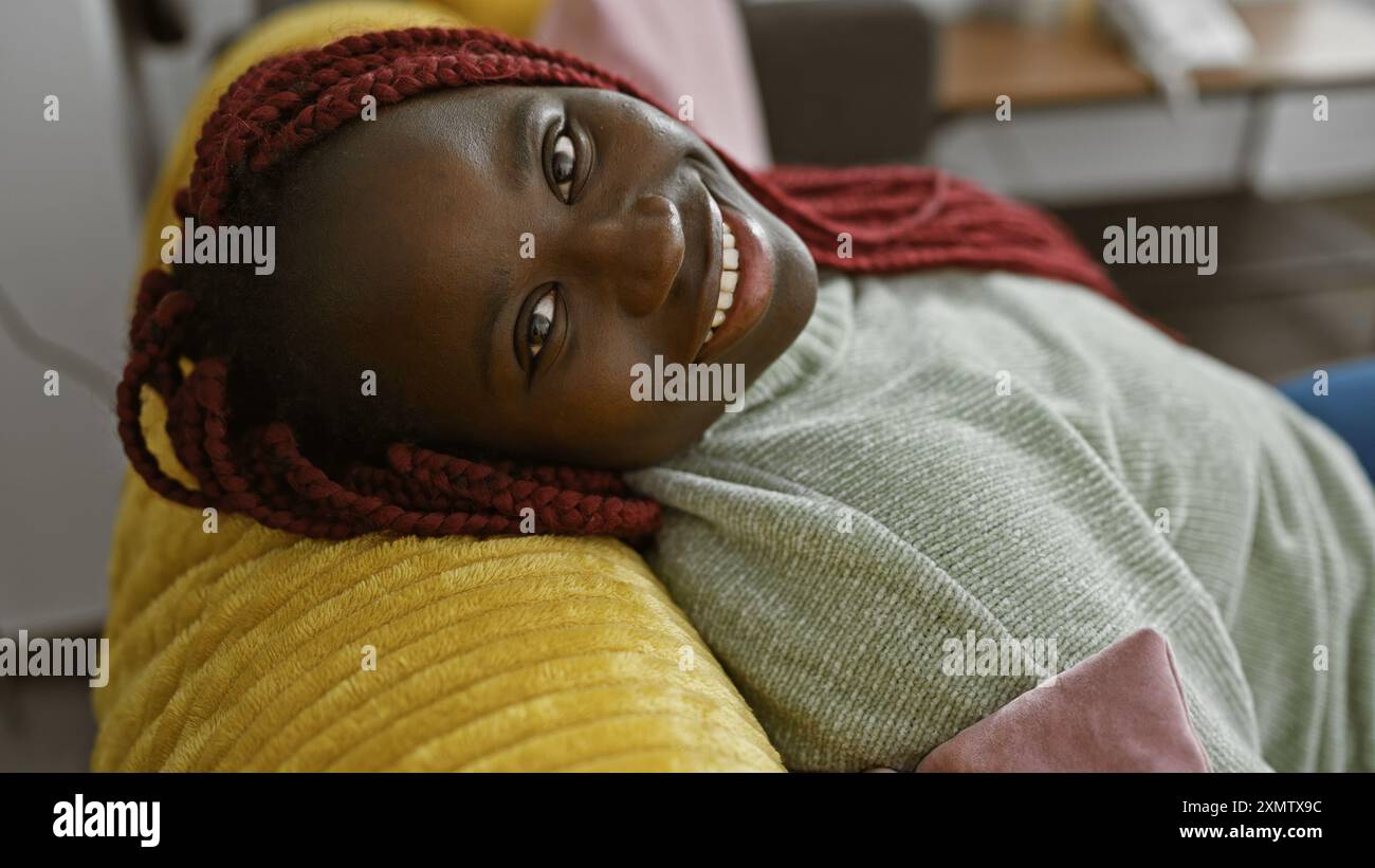 Eine lächelnde afroamerikanische Frau mit Zöpfen liegt bequem zu Hause auf einer gelben Couch und zeigt Schönheit und Entspannung. Stockfoto