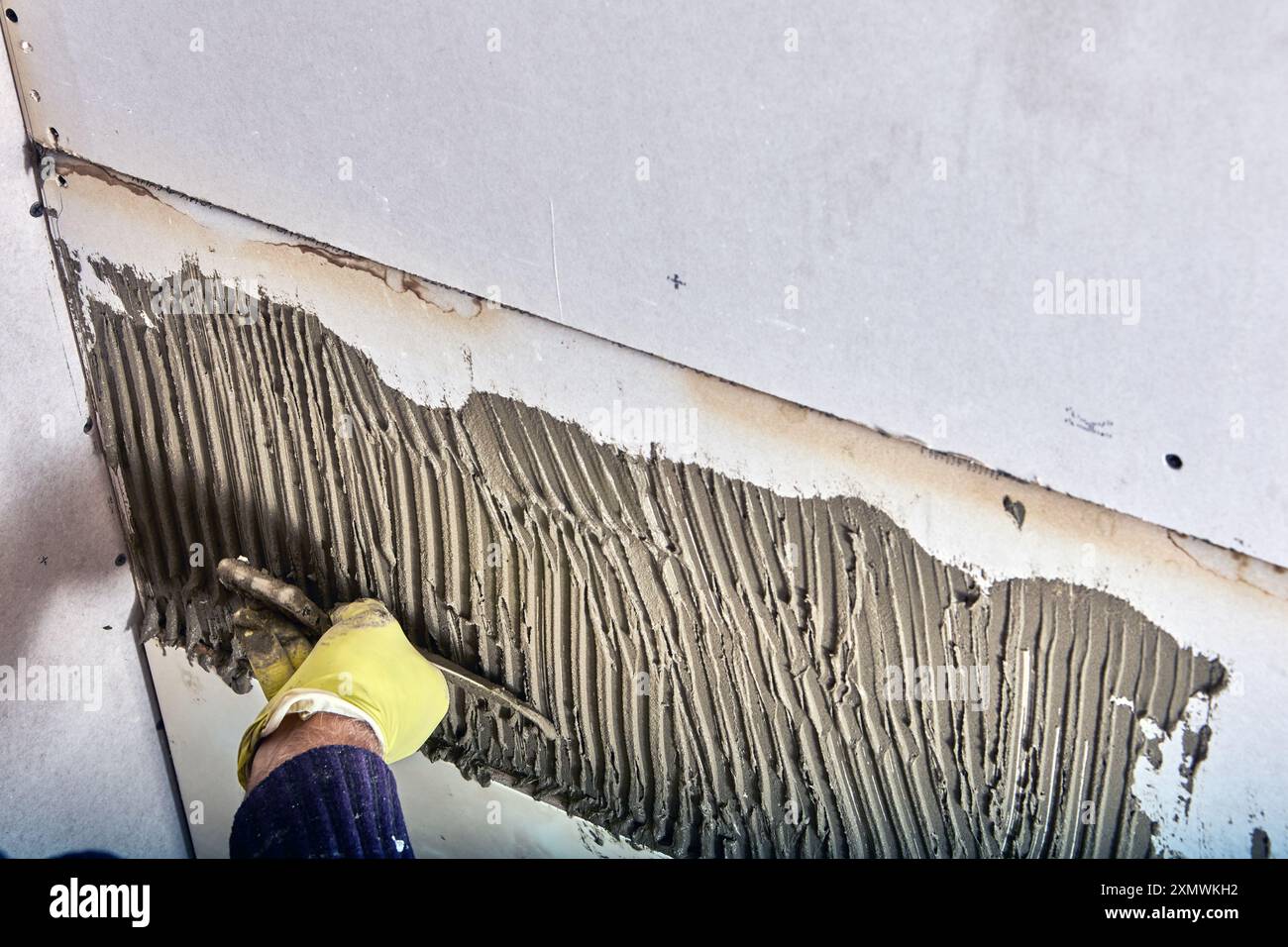 Die Fliesenmaschine des Bauarbeiters trägt mit einer gekerbten Kelle Klebermörtel auf die Wand auf. Stockfoto