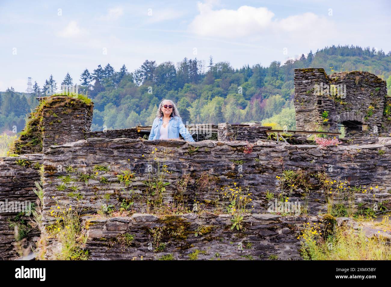 Seniorin, die zwischen Steinmauern steht, die von der Zeit getragen wurden, Besichtigungen der Burgruinen von La Roche-en-Ardenne, grüne Laubbäume auf Hi Stockfoto