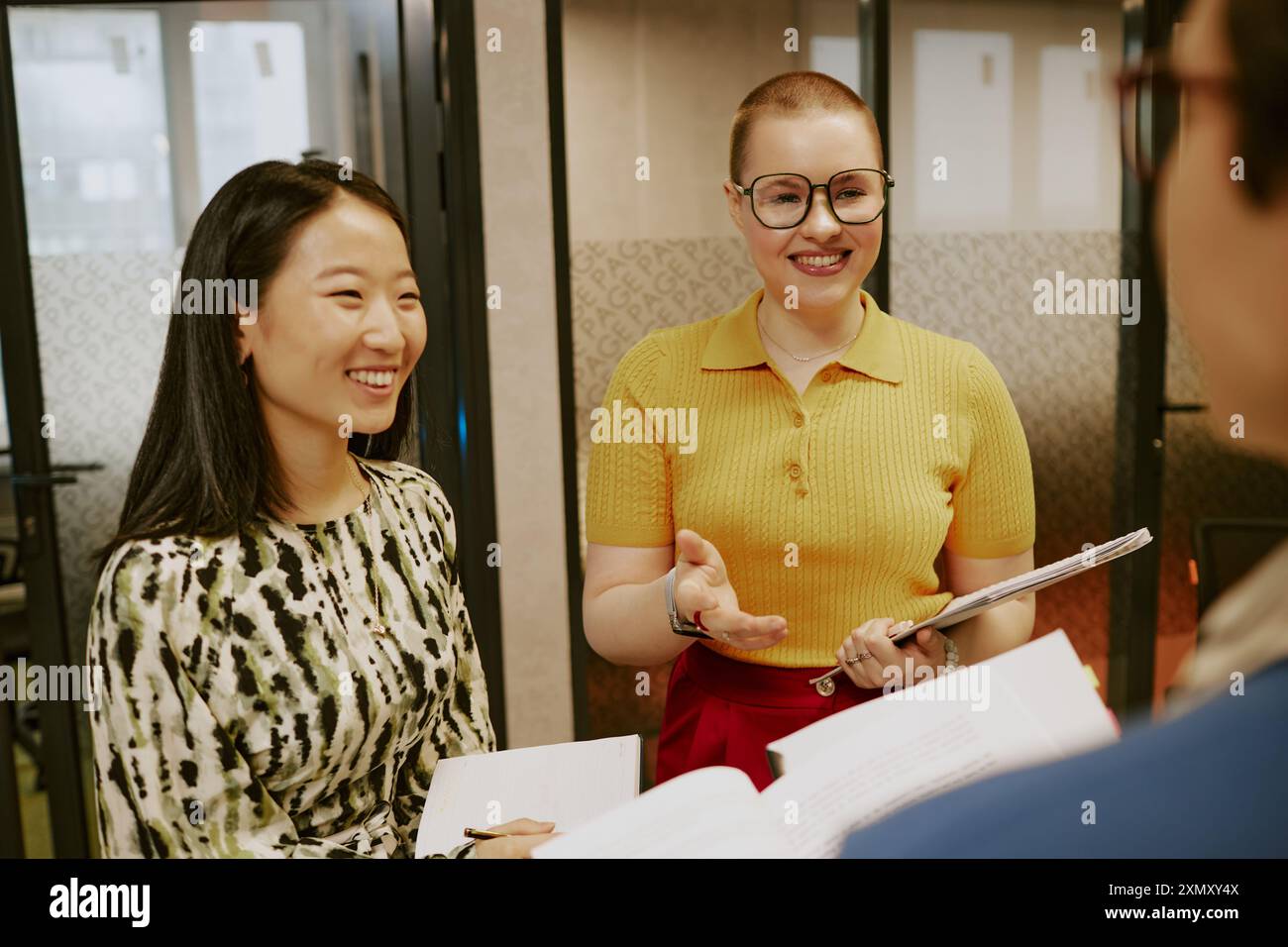 Zwei junge asiatische und kaukasische Managerinnen, die Notizbücher und Dokumente in einem modernen Büro halten und mit ihrer nicht erkennbaren Kollegin sprechen Stockfoto