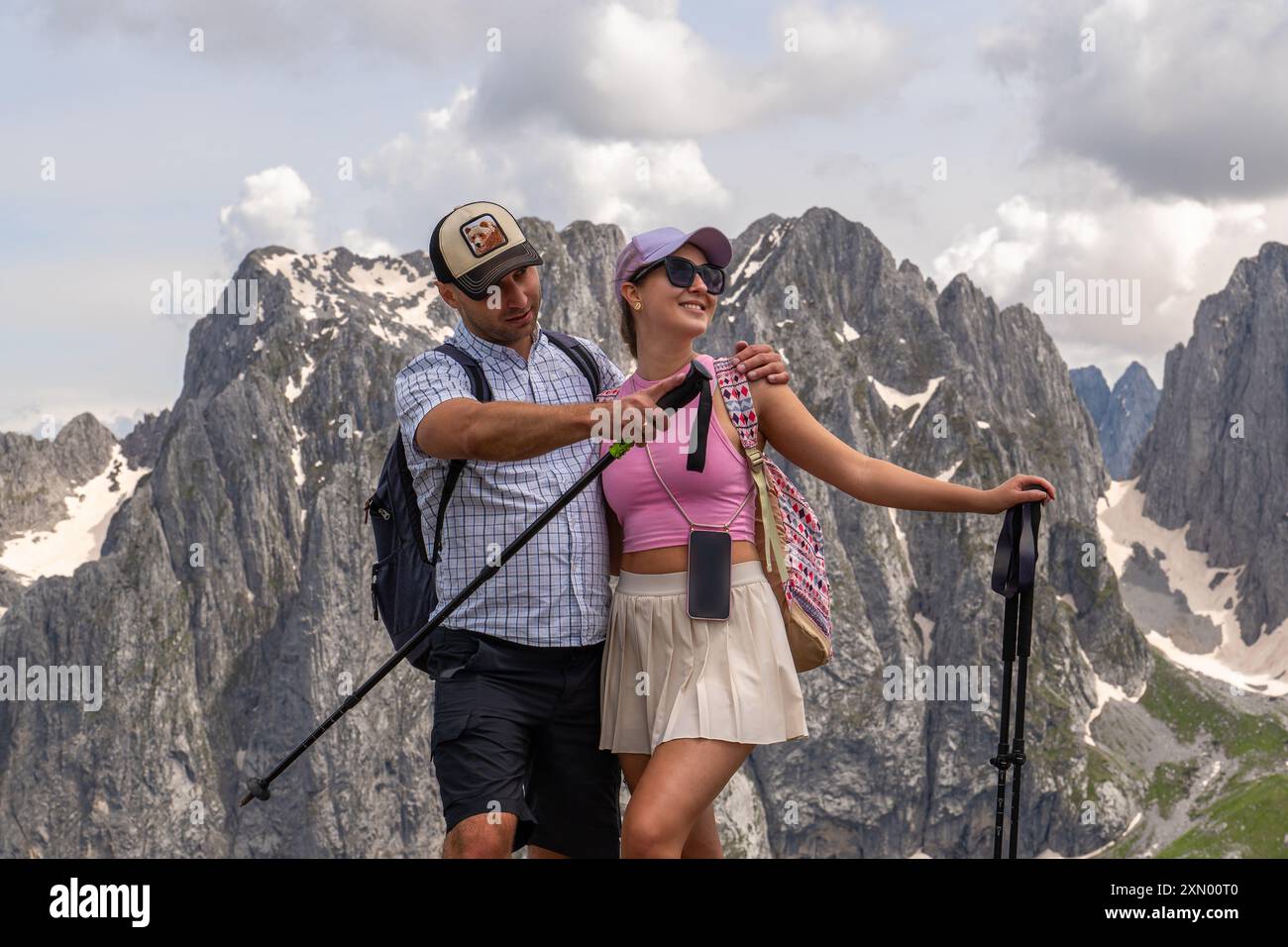 Ein junges Paar, ein Mann und eine Frau, die vor einem Hintergrund verschneite Berge stehen. Der Mann zeigt die Richtung, wohin er gehen soll. Sie lächeln und genießen es. Stockfoto